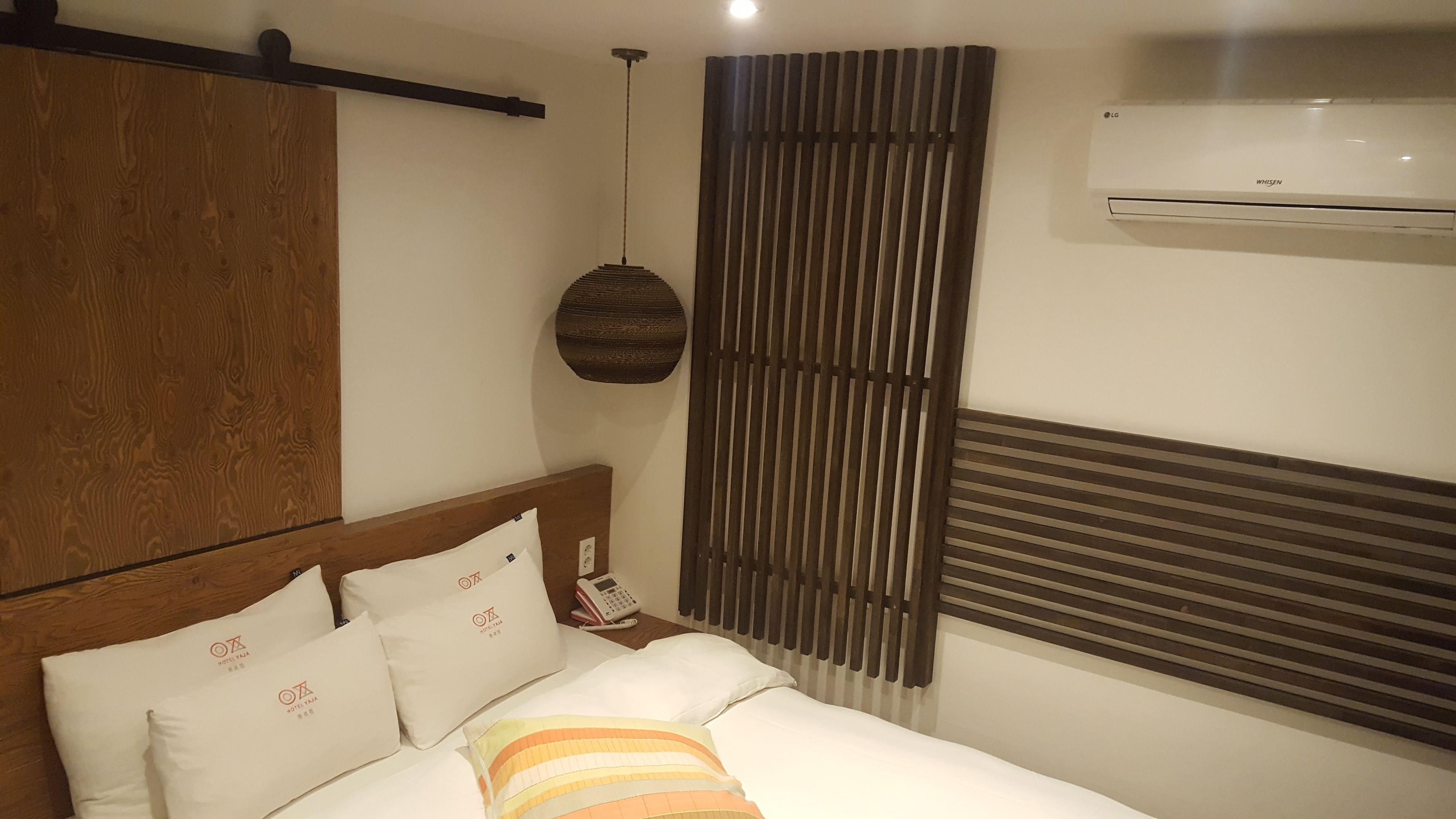 Habitación de hotel con cama, almohadas, teléfono, aire acondicionado de pared y lámpara colgante.