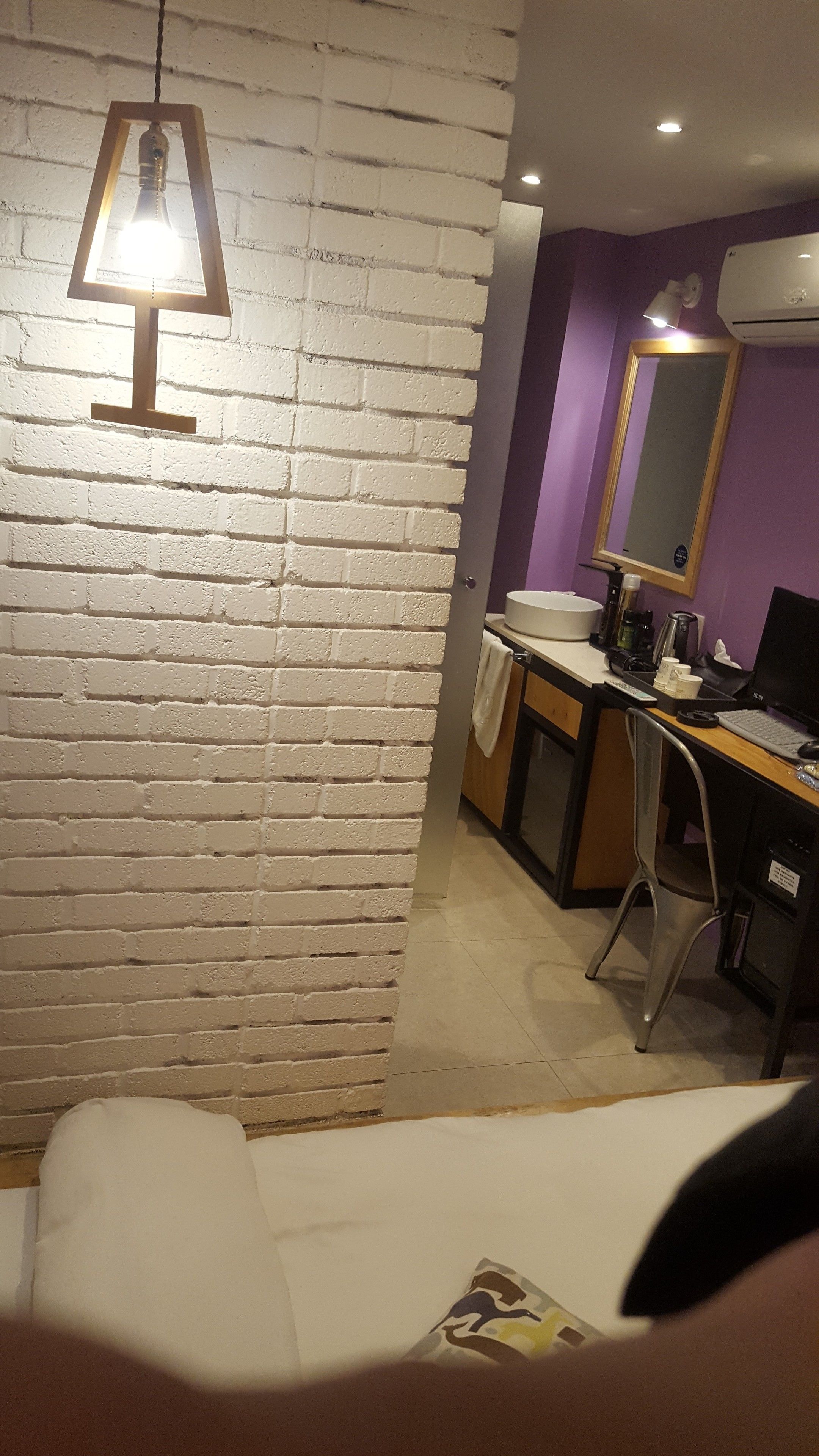 Een kamer met een bakstenen muur, een bureau met een computer, een stoel en een hangende lamp.