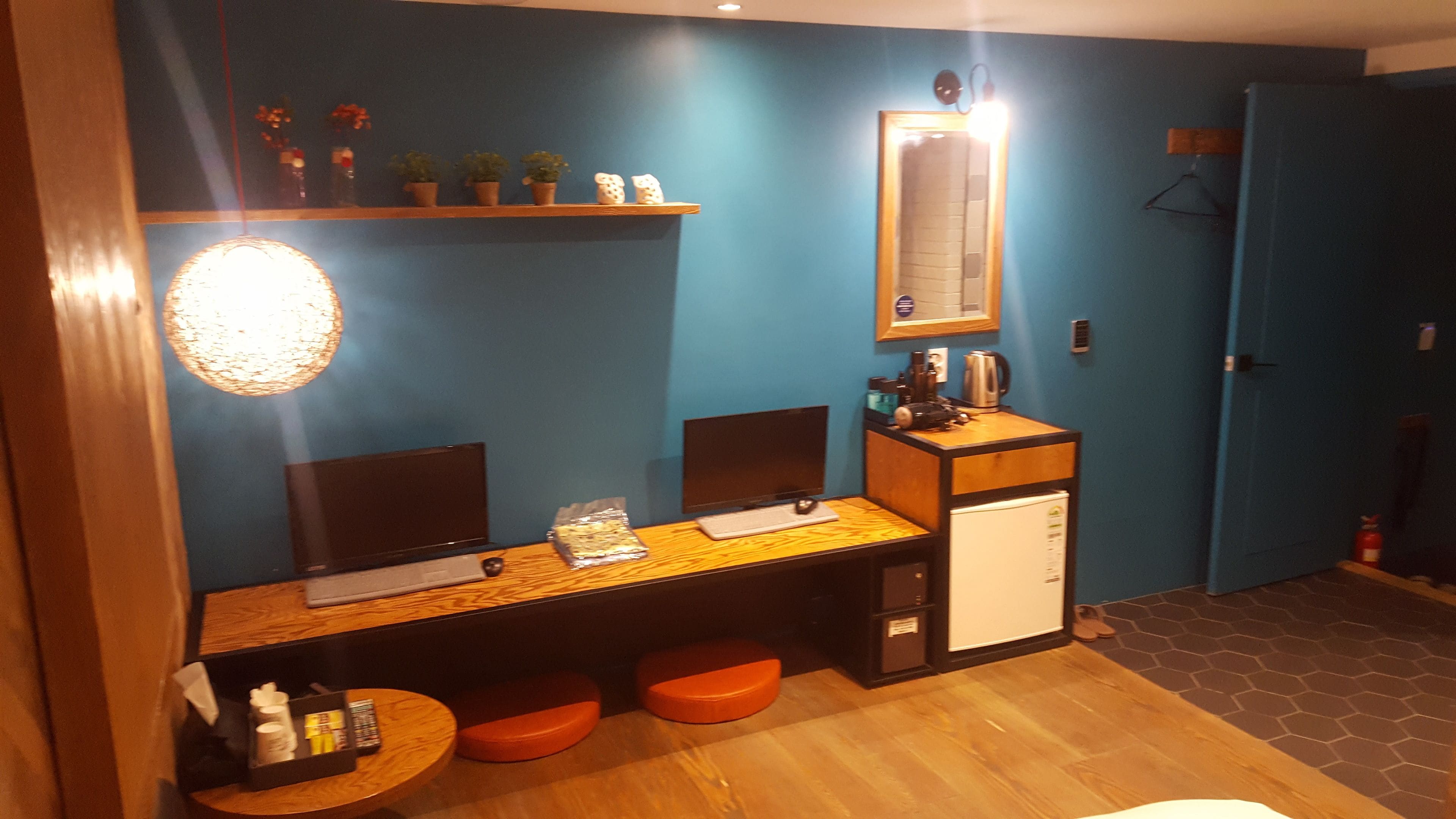 A room with a wooden desk, two monitors, a small refrigerator, and orange stools.