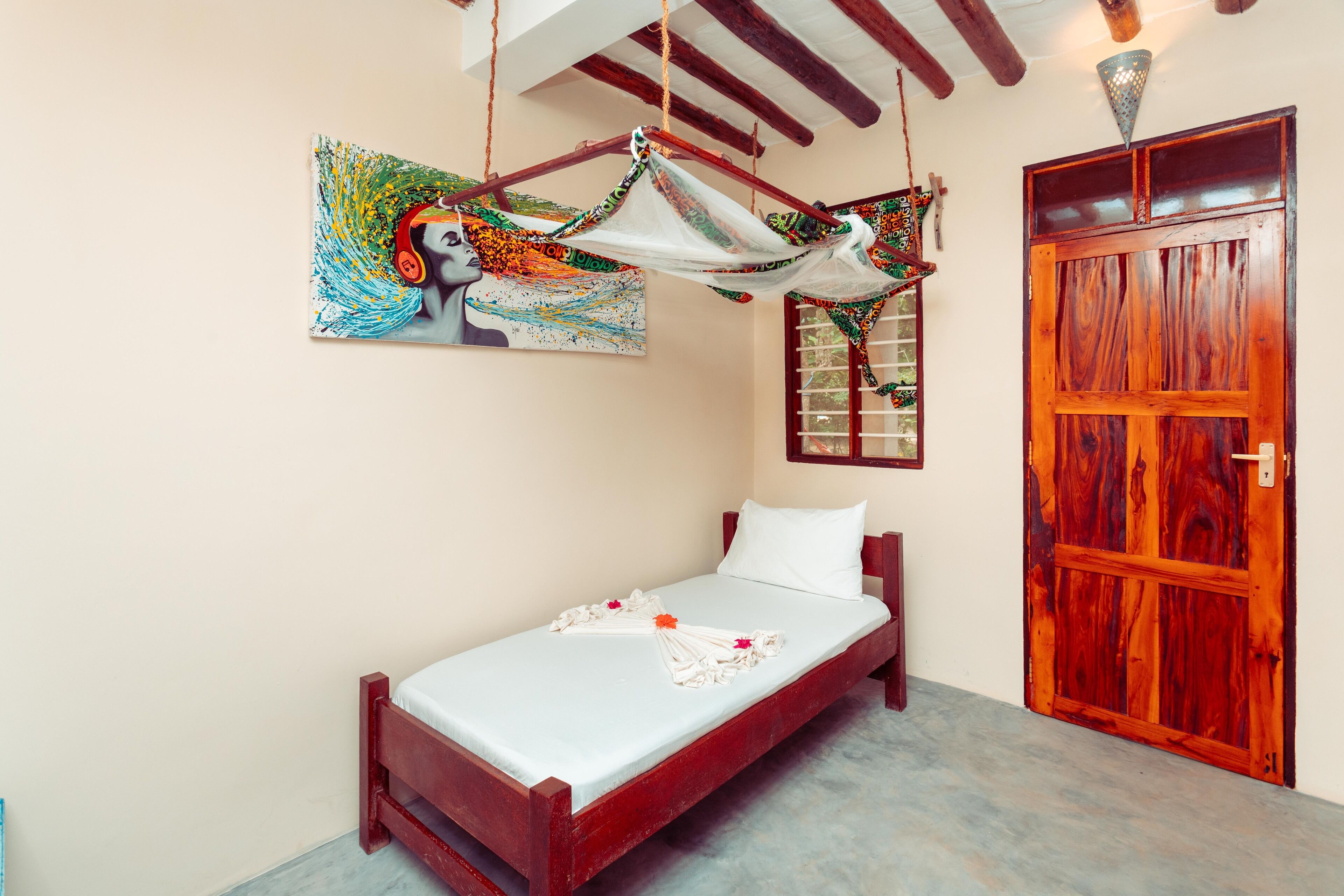 Una cama individual con colchón blanco y cabecero de madera, una puerta de madera y una hamaca colgada del techo.