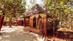 A rustic bungalow with a thatched roof, surrounded by dense foliage and a sandy ground. - Demani Lodge (Paje)