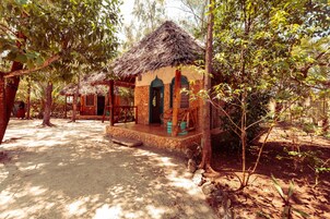 Ein rustikales Bungalow mit einem Reetdach, umgeben von dichtem Grün und sandigem Boden.