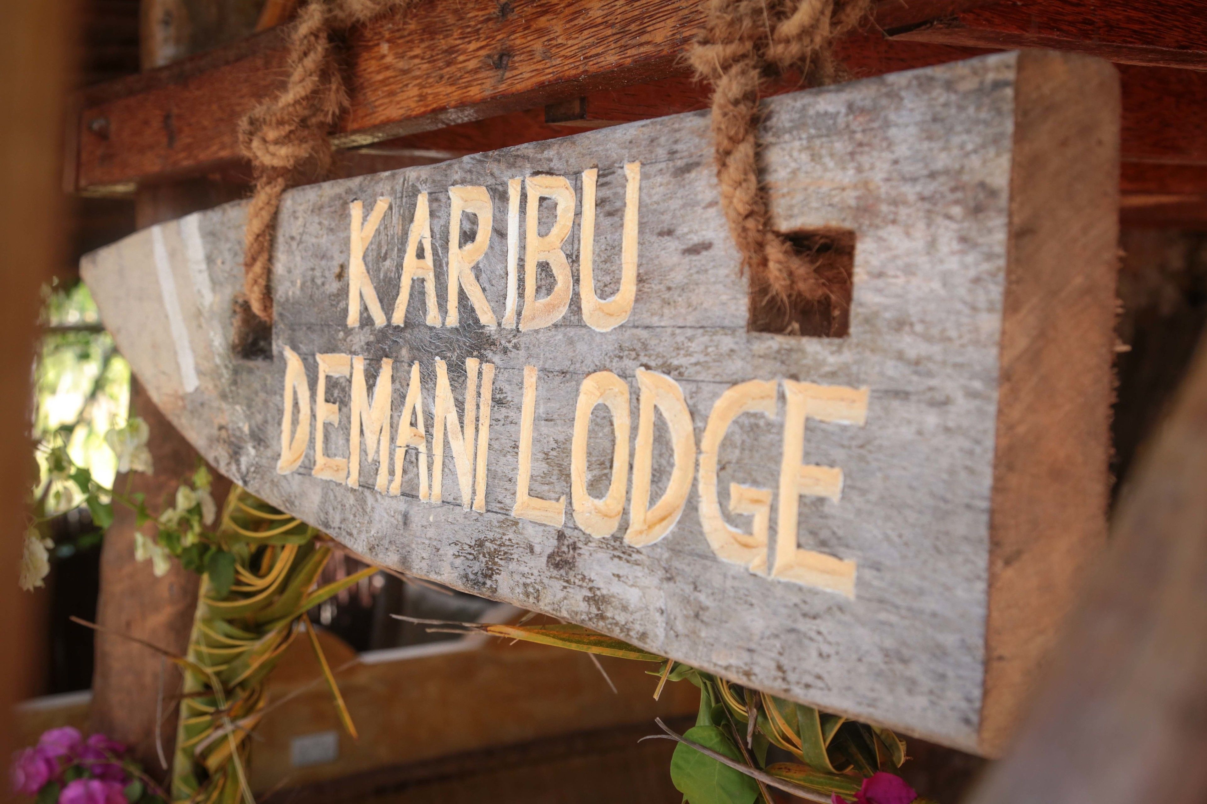 A wooden sign with the text "Karibu Deman Lodge" indicating the name of the lodge.