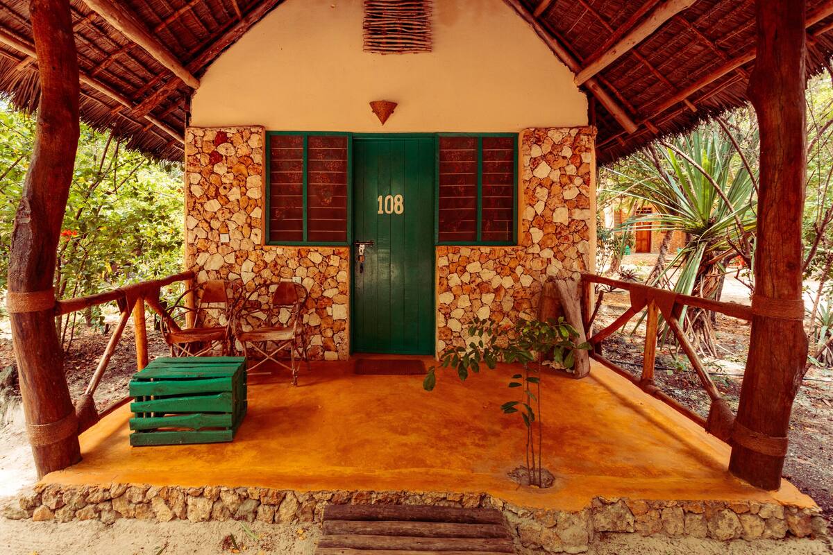 A rustic building with a green door marked "108", stone walls, wooden benches, and a thatched roof.