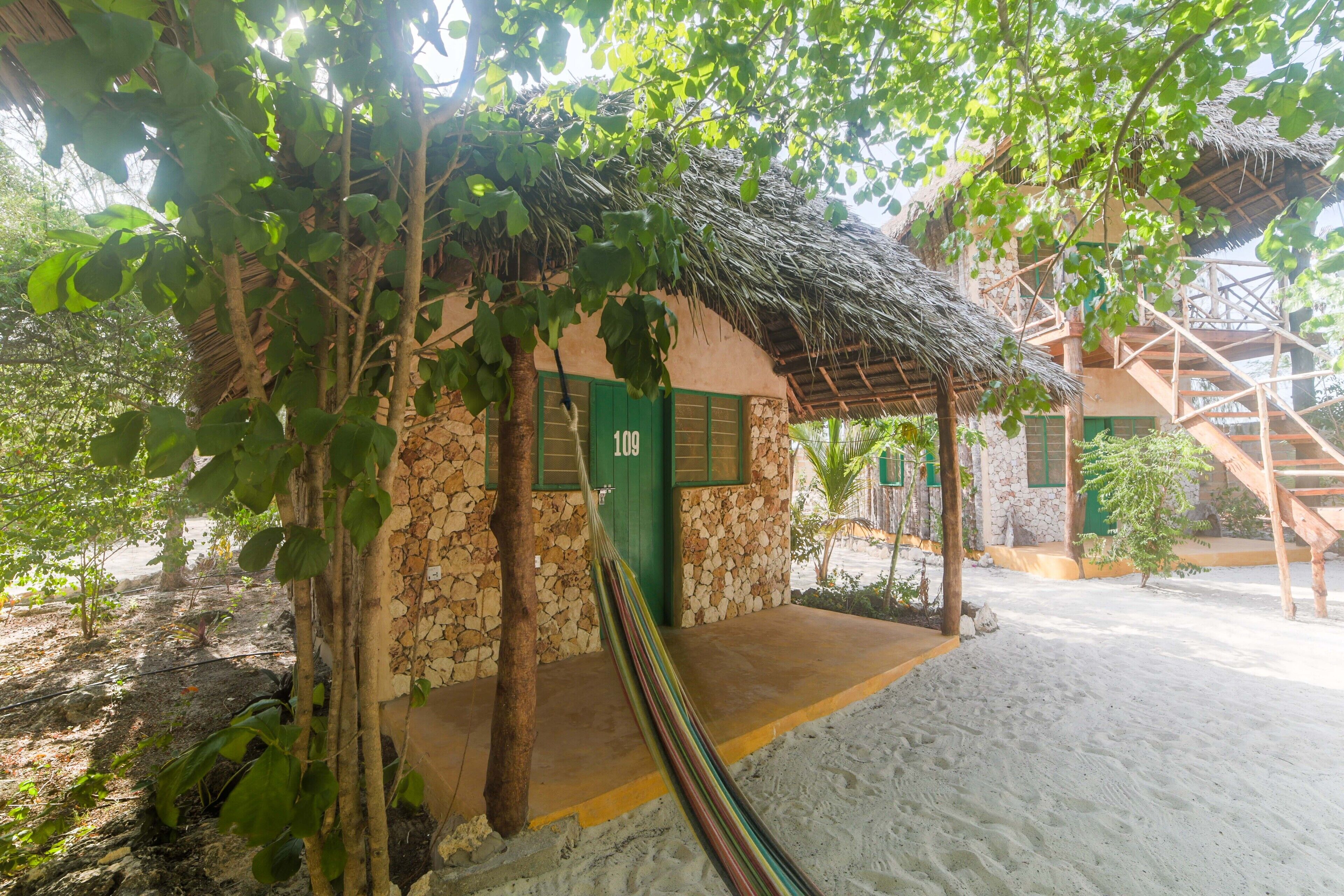A rustic building with a stone exterior and a thatched roof, a staircase leading to an upper level, and a hammock in the foreground.