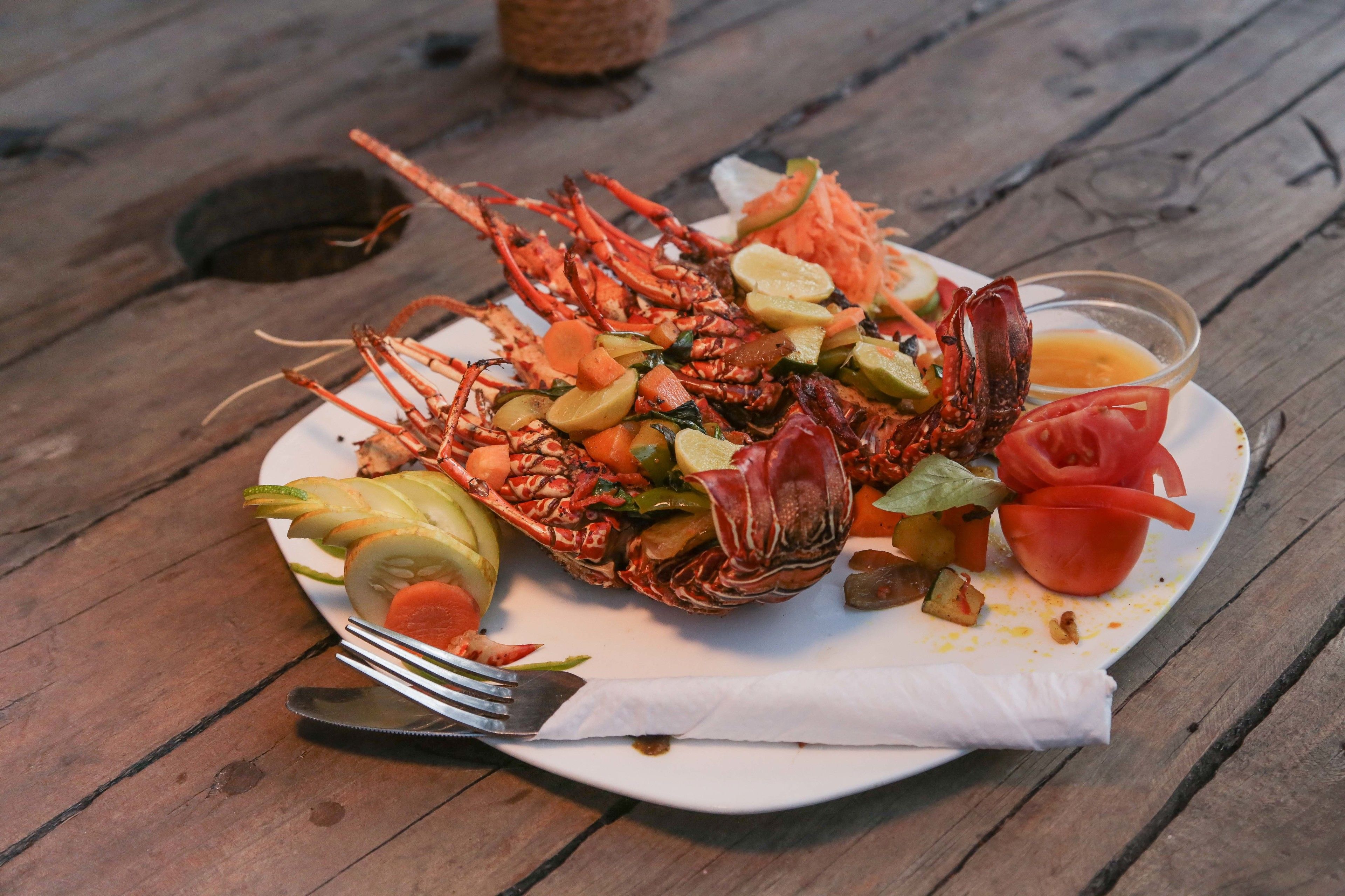A plate of cooked lobster with vegetables and a small bowl of sauce on a wooden table.