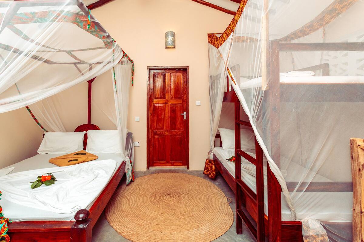 A room with two wooden beds, each with a canopy. There is a woven mat on the floor, a wooden door, and a wall-mounted light fixture.