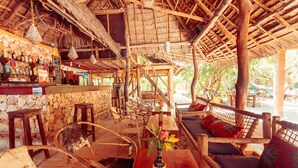 A rustic bar area with wooden furniture, a stone wall, and a thatched roof. - Demani Lodge (Paje)