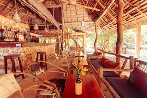 A rustic bar area with wooden furniture, a stone wall, and a thatched roof.