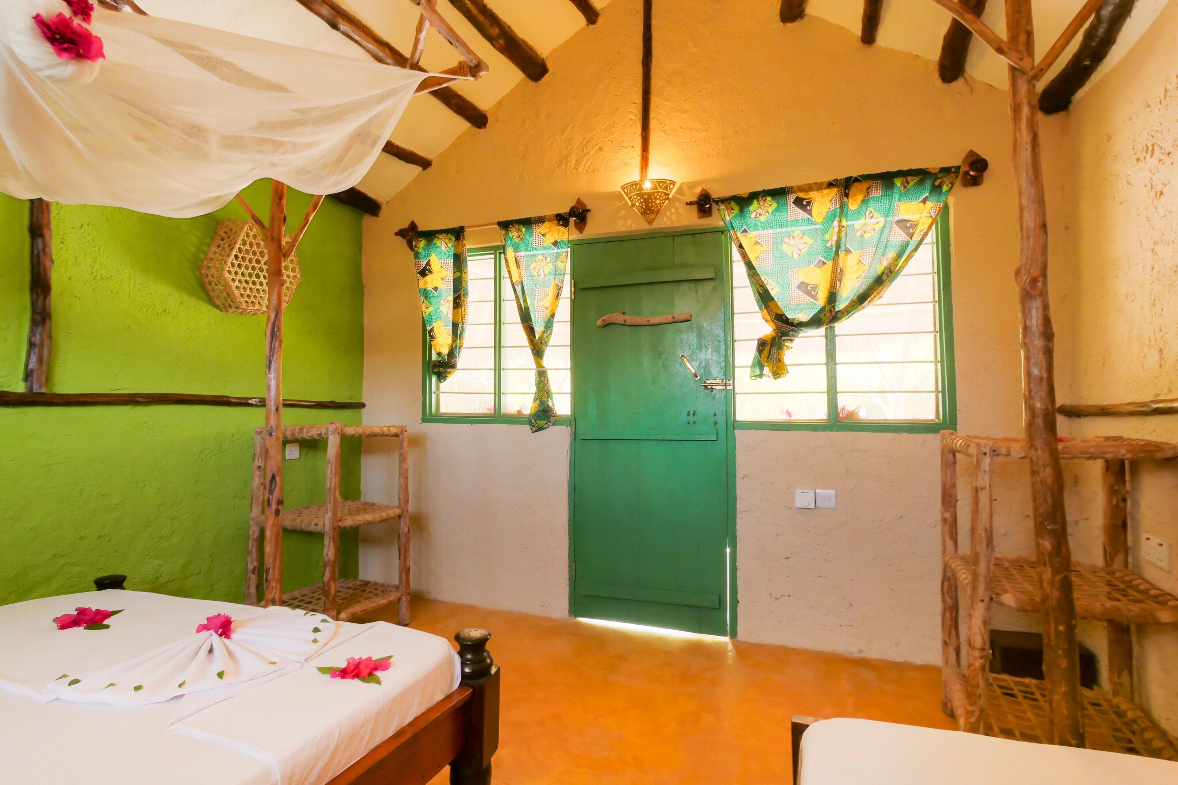 A room with a green door, a bed with a white bedspread and pink flowers, and a window with patterned curtains.