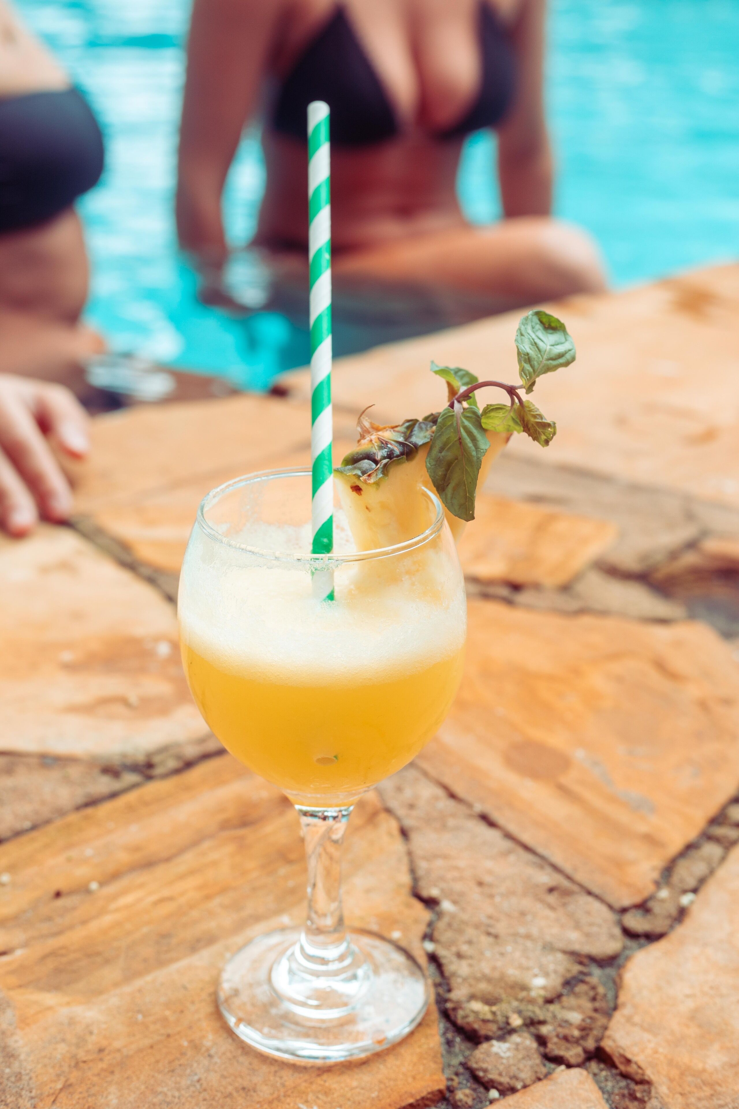 A cocktail with a green and white striped straw and a mint garnish on a wooden table near a pool.