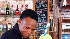 A person at a bar with a drink in front of them. There are bottles and a chalkboard menu in the background. - Demani Lodge (Paje)