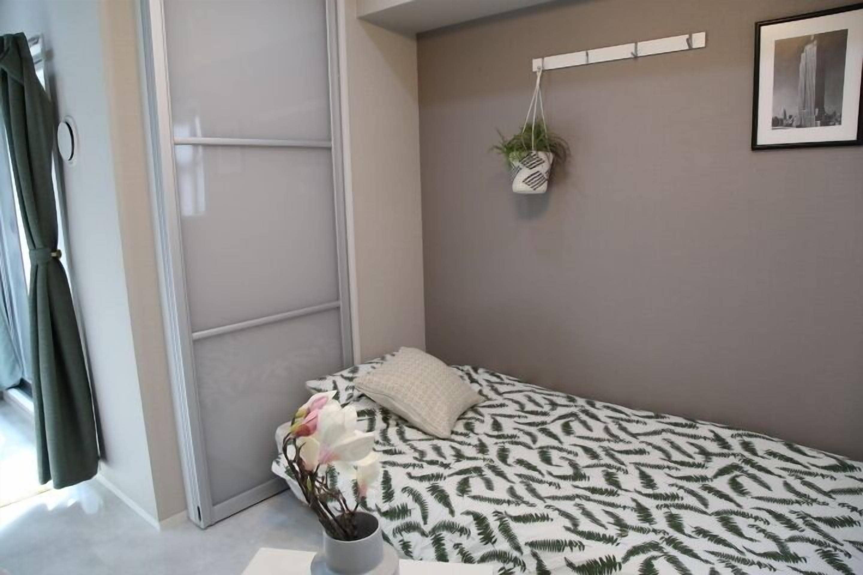 A small, neatly arranged bedroom with a bed, a bedside plant, and a framed picture on the wall.