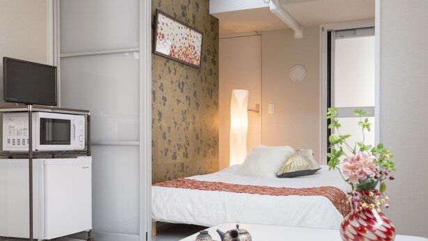 A hotel room with a bed, a bedside table with a vase and teapot, a microwave, and a TV on a cart. - Jesus Square Nipponbashi (Osaka)