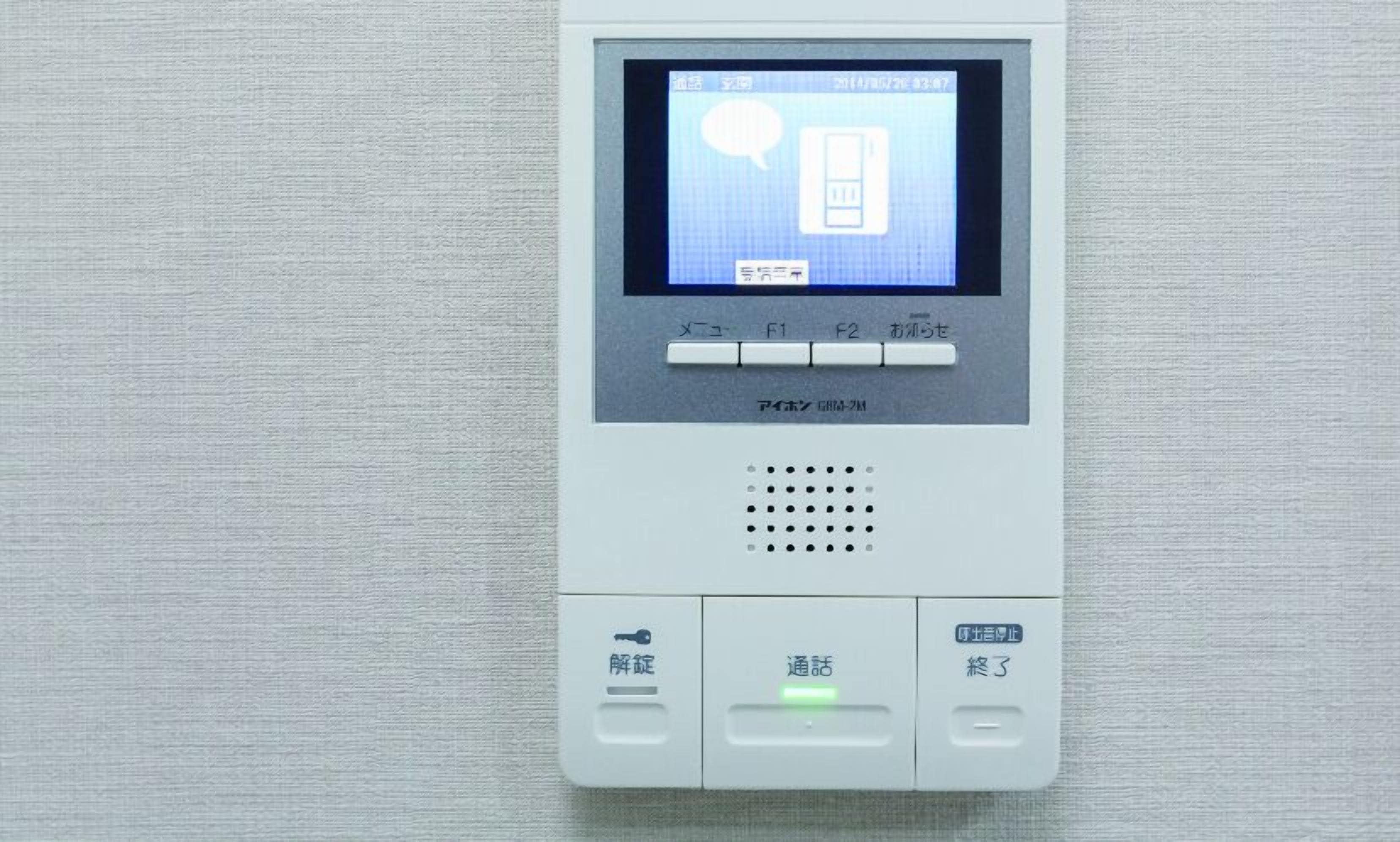 A digital intercom system with a screen displaying a door icon and Japanese text, a keypad, and a speaker.