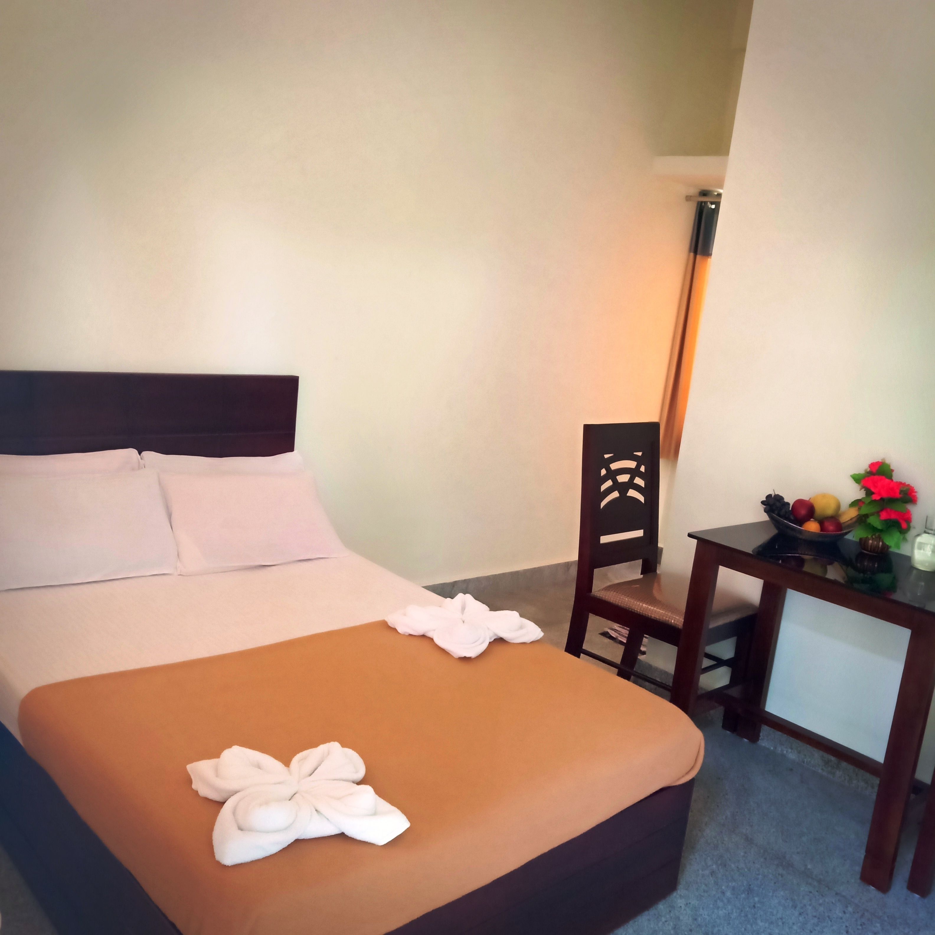 A hotel room with a bed, a chair, a small table with fruit and flowers, and a lamp.