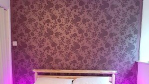 Un lit avec une literie blanche, dans une pièce dont le papier peint est à motifs floraux et qui est éclairée d’une lumière ambiante violette.