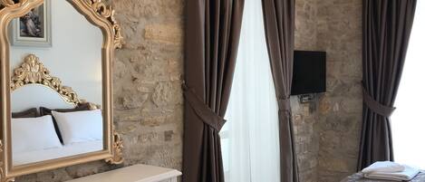 A bedroom with a stone wall, a large ornate mirror, a white console table, a black bench, a bed with a brown bedspread, and a chandelier.