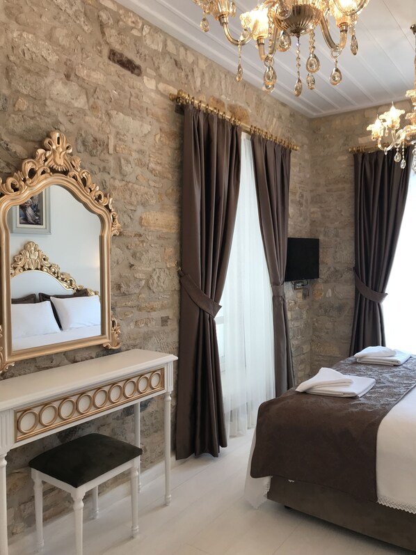 A bedroom with a stone wall, a large ornate mirror, a white console table, a black bench, a bed with a brown bedspread, and a chandelier.