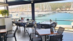 Zona de menjador interior amb taules i cadires, grans finestres amb vista a un port esportiu i embarcacions.