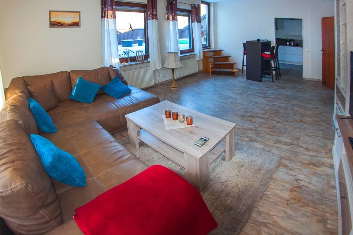 Un salón con un sofá de cuero marrón, una mesa de centro de madera y un otoman rojo. En el fondo, se observa una zona de cocina con una mesa de comedor y sillas.