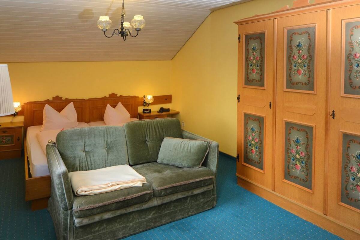 Una habitación con una cama, un sofá verde, un armario de madera con diseños florales y un candelabro.