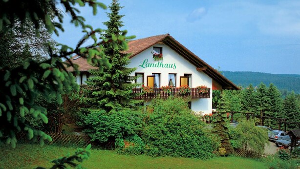 Un bâtiment traditionnel, de couleur blanche, avec un toit rouge et un balcon, entouré d’une végétation luxuriante et d’arbres.