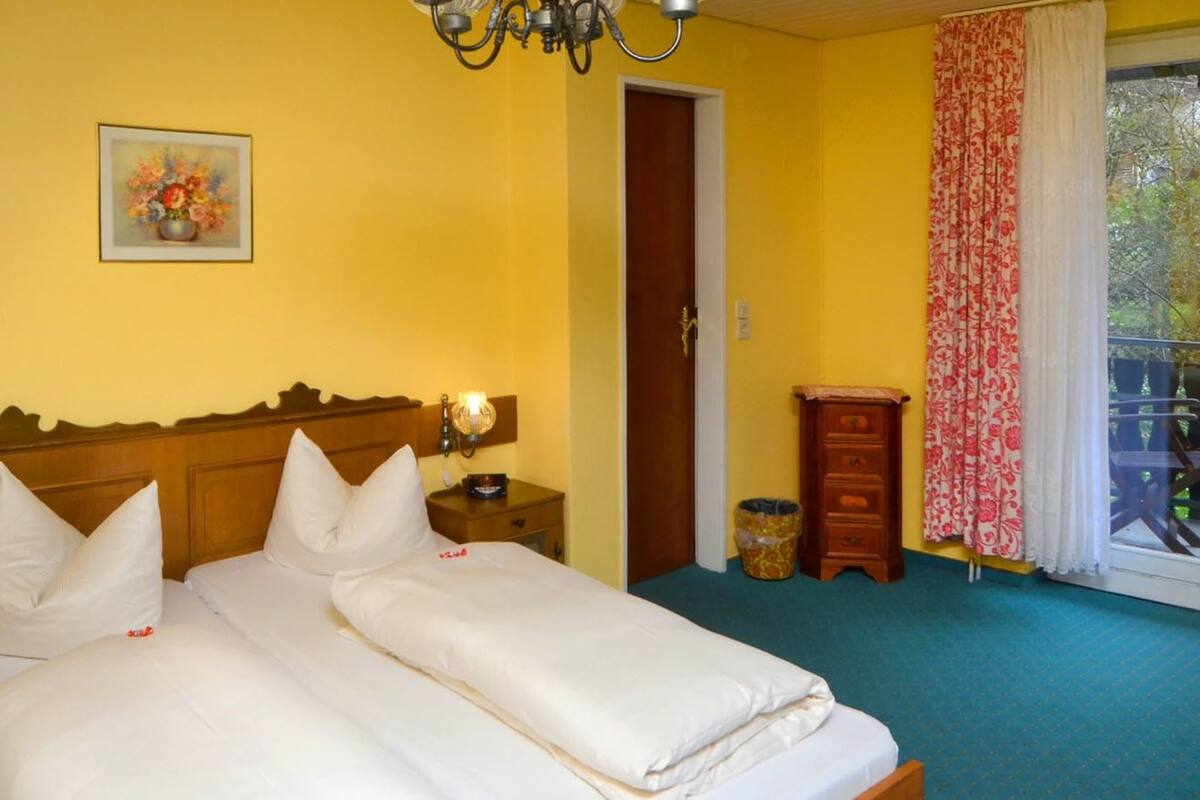 Habitación de hotel con dos camas, cabecero de madera, mesita de noche con lámpara, pequeño armario de madera, cesto de basura y ventana con cortina.