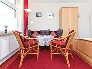 Um quarto com carpete vermelho, cadeiras de vime, uma pequena mesa com toalha branca e um guarda-roupa de madeira.