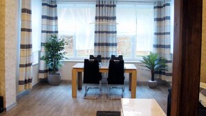 A dining area with a table and chairs, large windows with striped curtains, and potted plants. - Ferienwohnungen Villa Waldblick (Zempin)