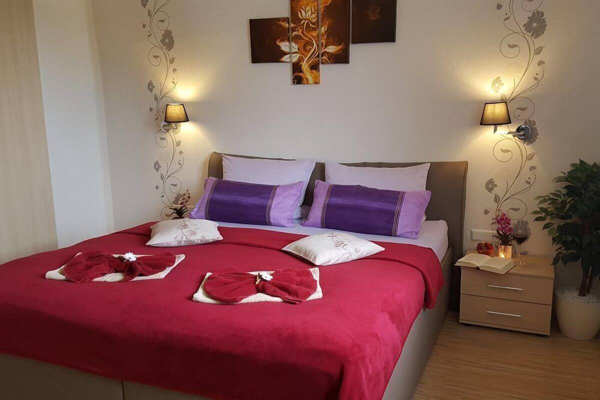 A neatly made bed with red and white linens, two wall-mounted lamps, and a small bedside table with a plant.