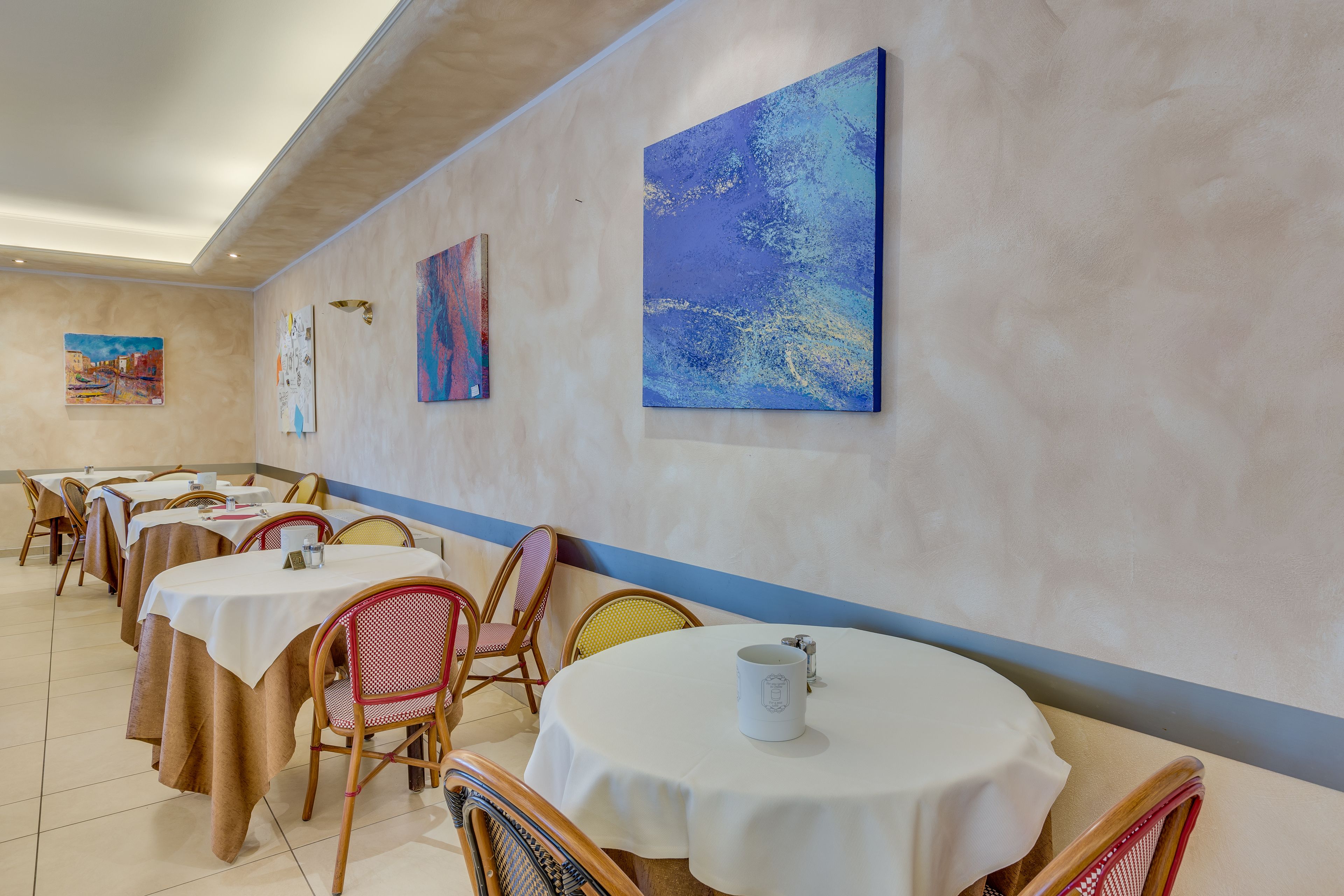 A dining area with round tables and chairs, artwork on the walls, and a blue stripe on the ceiling.
