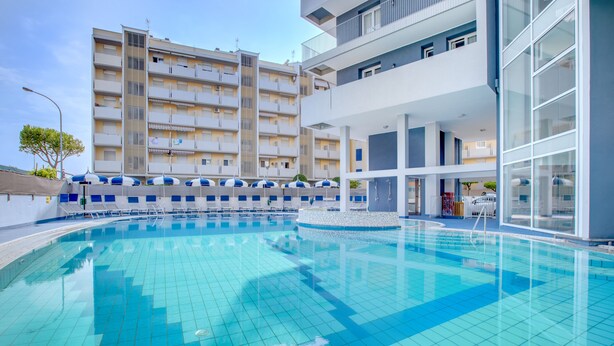 A swimming pool with tiled flooring, a covered area with umbrellas, and a multi-story building with balconies in the background. - Hotel Ambassador (Caorle)