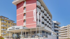 A modern hotel with a red and white facade, multiple balconies, and a prominent entrance with a sign. - Hotel Ambassador (Caorle)