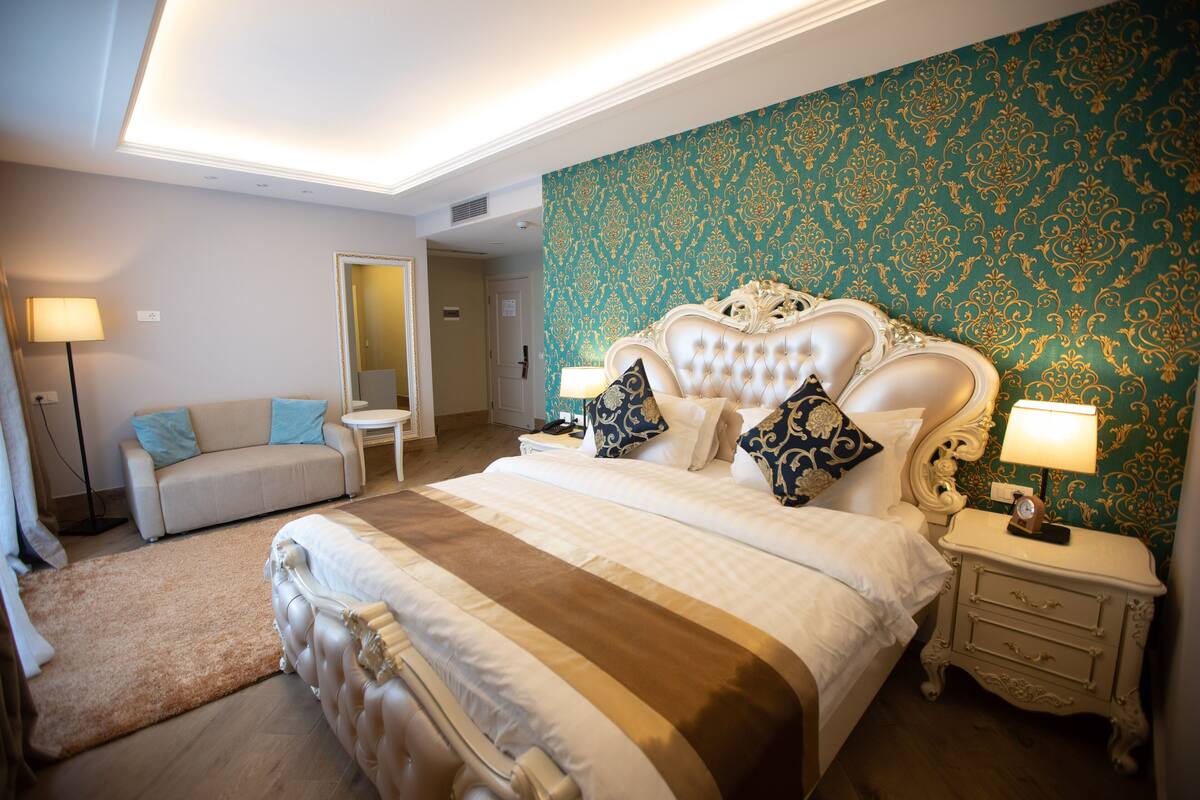 A bedroom with a large bed, a nightstand, a bedside table, a lamp, a sofa, and a mirror.