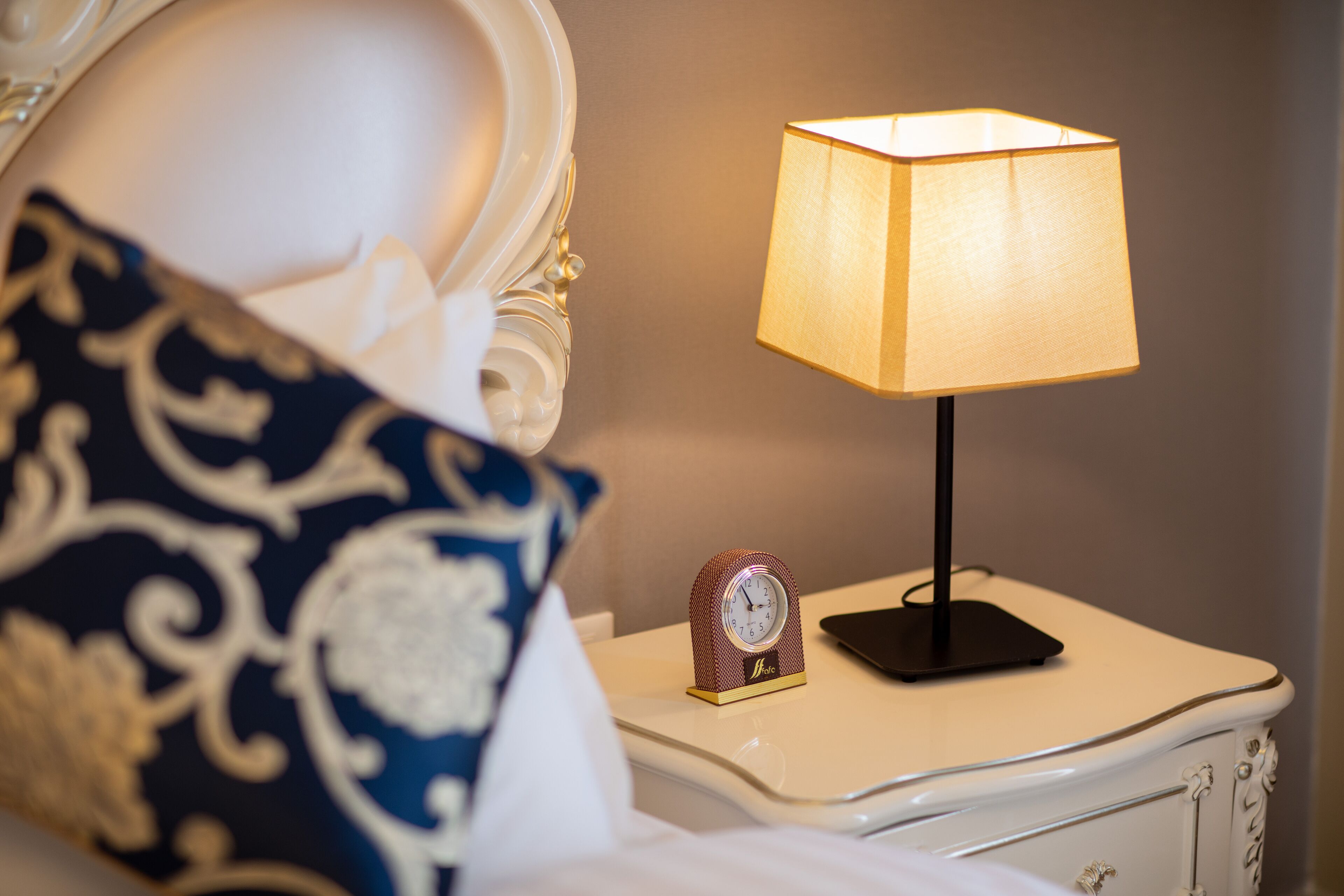 Ein ordentlich bezogenes Bett mit einem gemusterten Kopfkissen, ein Nachttisch mit Lampe und eine kleine Uhr.