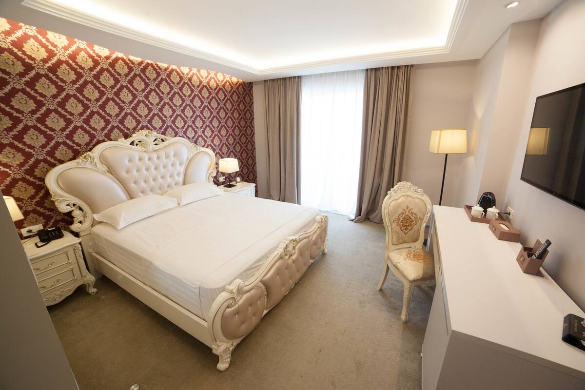 A hotel room with a large bed, a desk, a chair, a TV, and a patterned wall.