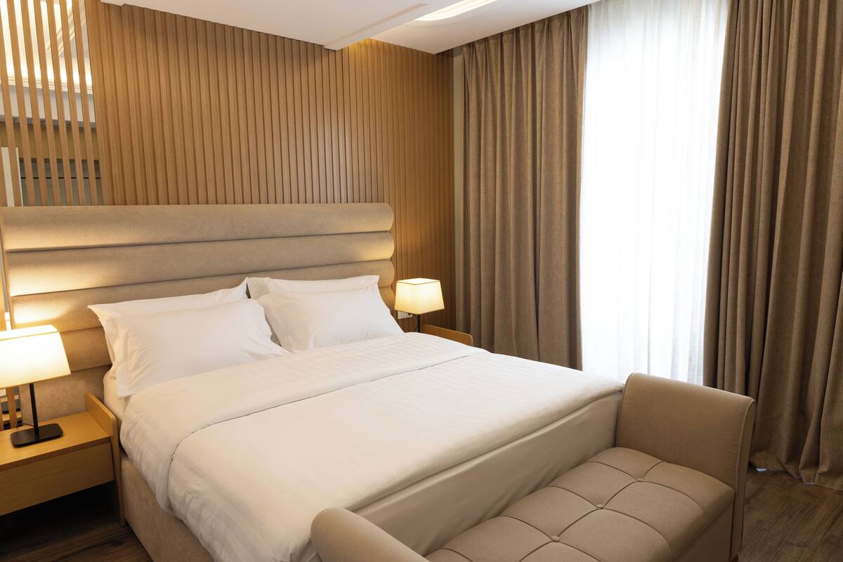 A hotel room with a large bed, two bedside tables with lamps, a headboard, and a view of the outside through curtains.