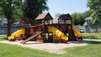 A playground with multiple slides and climbing areas, surrounded by grass and trees.