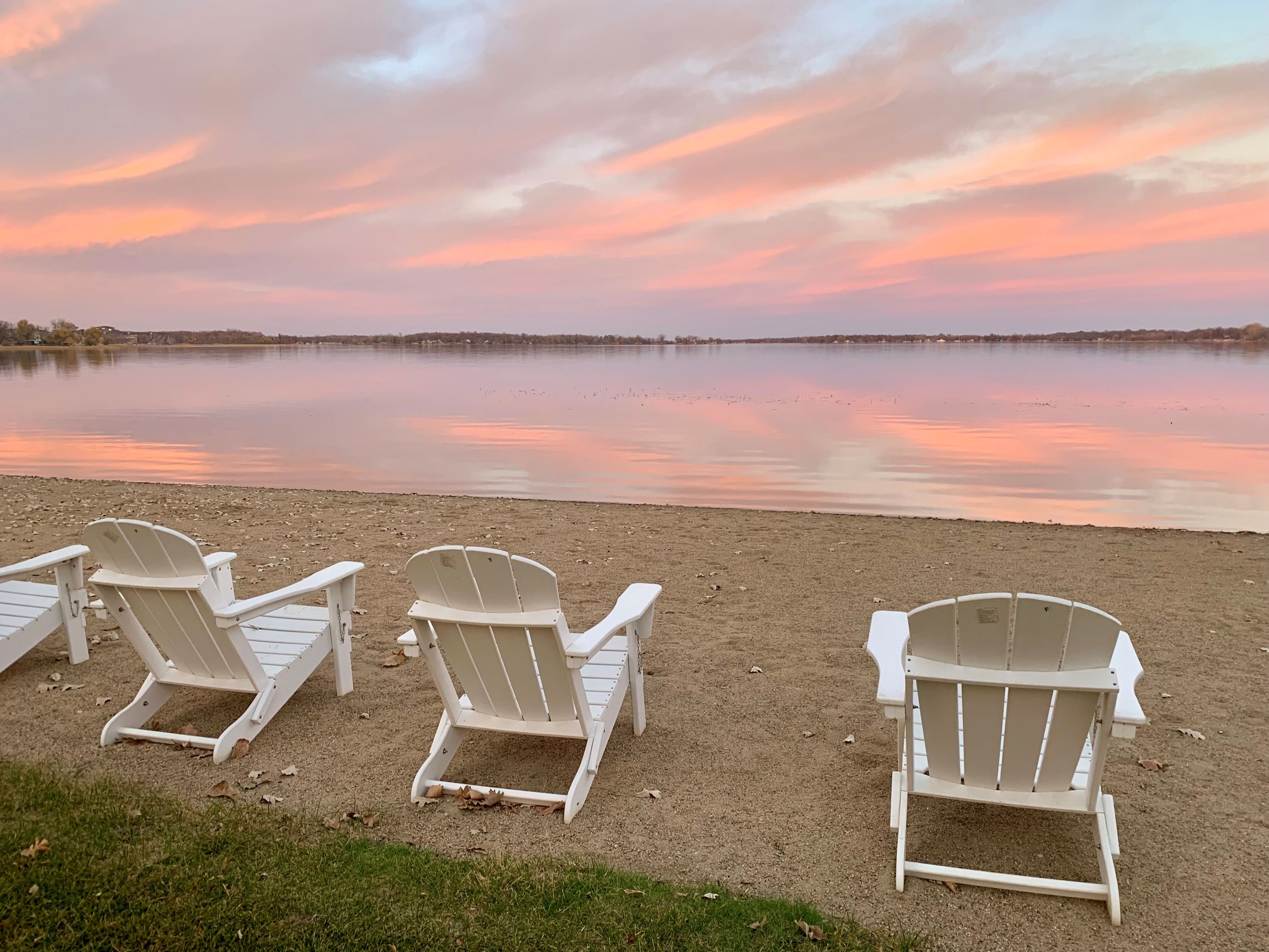 Three white Adirondack chairs on a sandy beach with a calm lake and a sunset in the background.