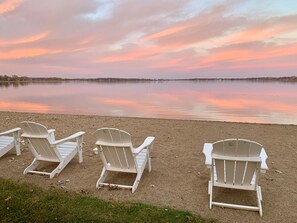 Three white Adirondack chairs on a sandy beach with a calm lake and a sunset in the background.