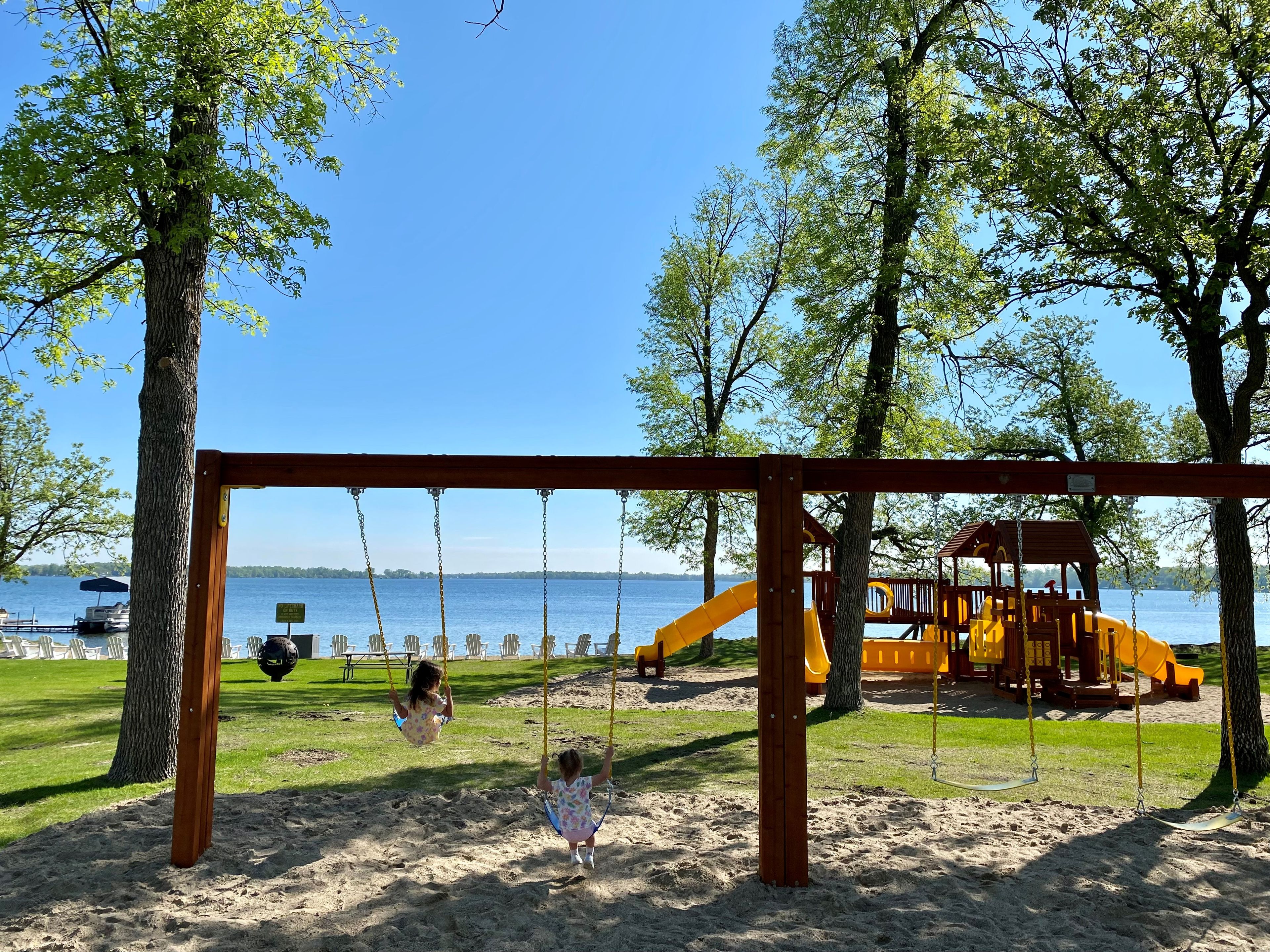 A playground with swings and a slide, set against a backdrop of trees and a body of water.