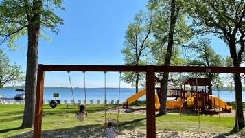A playground with swings and a slide, set against a backdrop of trees and a body of water.