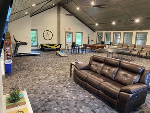 Un grand espace intérieur comprenant un canapé en cuir marron, une table de billard, une table de ping-pong, un tapis de course et une table à manger avec des chaises.
