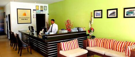 A hotel reception area with a reception desk, chairs, a sofa set, and framed pictures on the wall.