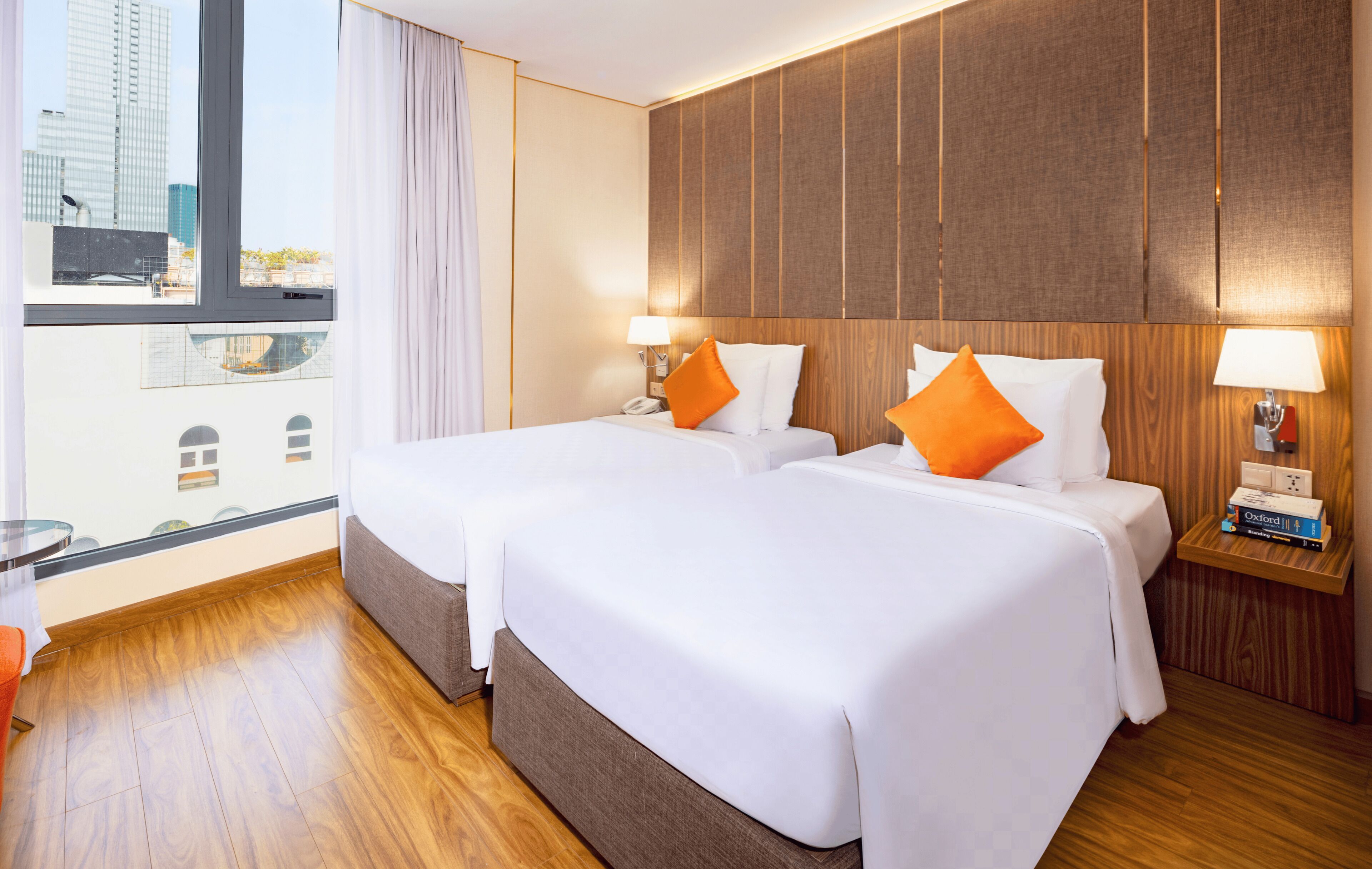 A hotel room with two beds, wooden paneling on the walls, a city view, and a small table with a lamp and books.