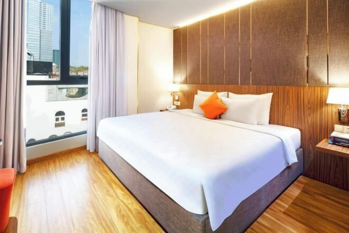 A hotel room with a large bed, wooden headboard, a nightstand, and a view of buildings through a window.