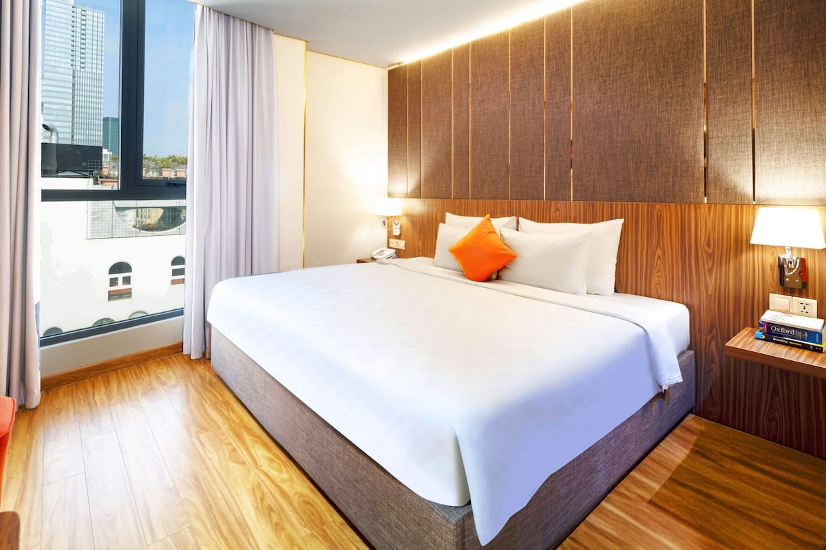 A hotel room with a large bed, wooden headboard, two bedside tables with lamps, a red chair, and a view of the city through a large window.