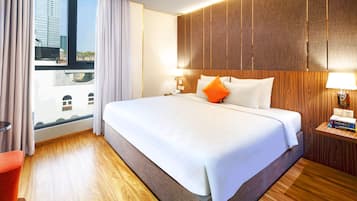 Ett hotellrum med en stor säng, ett träbeklätt sänggavel, två sängbord med lampor, en röd stol och utsikt över staden genom ett stort fönster.