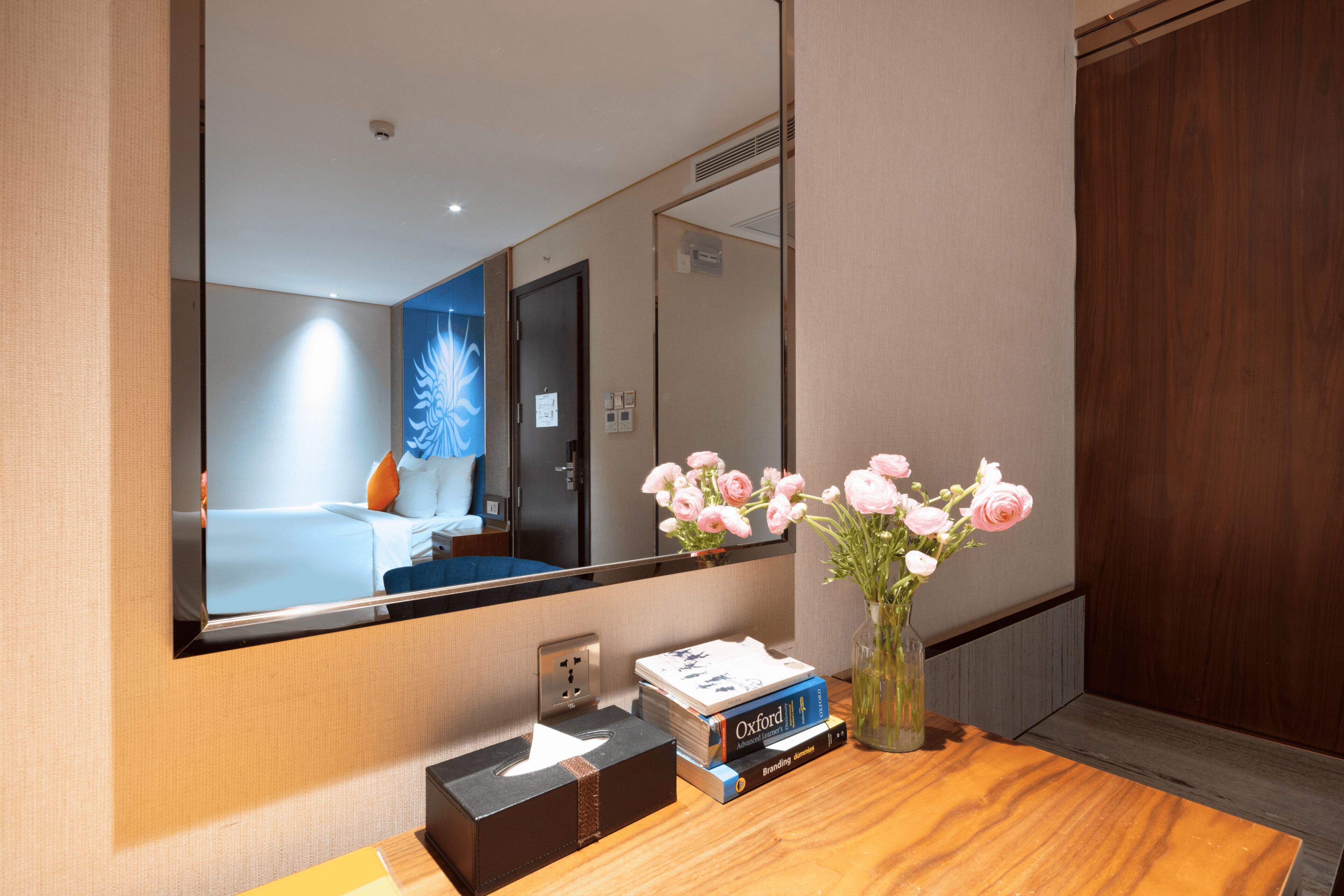 A hotel room with a wooden desk, a mirror reflecting the room, a vase with flowers, and a stack of books.
